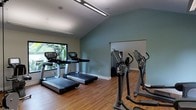 This fitness center is a thoughtfully designed single-level exercise facility built to support a comprehensive cardio workout experience. The room features vaulted ceilings that lend an open, airy feel, while soft-green accent walls create a calm and motivating atmosphere throughout the space.

The facility is outfitted with a row of modern treadmills positioned directly in front of large windows, offering unobstructed views of lush, tropical greenery outside — providing a natural and energizing backdrop for workouts. Alongside the treadmills, a bank of elliptical trainers and stair-climber machines lines the opposite side of the room, ensuring a well-rounded selection of cardio equipment for users of varying fitness levels.

Warm hardwood-style flooring runs throughout the space, adding a clean and polished aesthetic that contrasts nicely with the cool-toned walls. Overhead, linear fluorescent lighting ensures the room is evenly and brightly illuminated, complementing the natural light that filters in through the large windows. The layout is open and uncluttered, allowing for comfortable movement between machines. Overall, this fitness center balances functionality with an inviting design, making it a practical and pleasant environment for daily exercise.