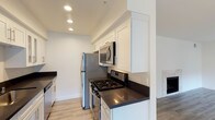 This well-appointed single-floor residence offers a clean, modern aesthetic highlighted by light wood-look flooring and crisp white walls that run consistently throughout the space. The galley-style kitchen is a functional standout, featuring white shaker cabinetry, dark countertops, and a full suite of stainless steel appliances including a gas range, over-the-range microwave, refrigerator, and dishwasher, all illuminated by recessed lighting. The open living area is generously sized, anchored by a tile-surround fireplace that serves as a focal point, with ample room for varied furniture arrangements. A separate area with large windows along one wall allows abundant natural light to fill the space, with views of greenery outside. At least one bedroom benefits from sliding glass doors that open directly to a private balcony, providing outdoor access and additional natural light. A second bedroom features a large window with a roller shade. The two bathrooms mirror each other in finish quality, each offering white cabinetry, dark countertops, and a bathtub-shower combination enclosed by sliding frameless glass doors. The overall design is cohesive and contemporary, emphasizing light, openness, and practical livability throughout.