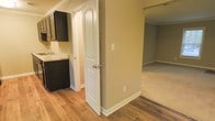 This single-story space is designed with a practical and cohesive layout that connects its key areas seamlessly. The hallway serves as a central corridor, featuring warm wood-look flooring that extends throughout the main areas, complemented by neutral beige walls and clean white baseboards that create a bright, understated aesthetic.

To one side, the kitchen is outfitted with dark cabinetry that provides a strong visual contrast against the lighter walls and countertops. A sink with a light-toned countertop is visible, and the dark cabinet finish adds a contemporary touch to the functional space.

A white interior door with an arched panel detail introduces a subtle architectural accent, adding character to what might otherwise be a utilitarian corridor. The door's classic paneled design is a notable design element within the space.

Adjacent to the hallway, a carpeted room is visible through an open doorway, suggesting a bedroom or secondary living area. The carpet offers a softer, quieter surface in contrast to the hard flooring found elsewhere. Overall, the space presents a clean, move-in ready environment with consistent finishes and a straightforward, functional floor plan.