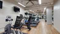 This single-floor amenity space comprises two distinct areas: a fully equipped fitness center and a dedicated laundry room, designed to serve the practical and wellness needs of its users. The fitness center is a generously sized, open workout space featuring light wood flooring and painted concrete block walls that give it a clean, functional aesthetic. The room is outfitted with a robust selection of cardio equipment, including multiple treadmills from brands such as True and Star Trac, stationary bikes, elliptical trainers, and a rowing machine. Ceiling fans circulate air throughout the space, while rows of fluorescent lighting and wall-mounted flat-screen televisions enhance the workout experience. Full-length mirrors line entire walls, amplifying the sense of space and allowing users to monitor their form. Exercise balls and free weights are also present, rounding out the available fitness options. The laundry facility is a bright, efficiently organized room finished with light tile flooring and a drop ceiling with recessed panel lighting. It houses a generous number of top-load washing machines and stacked dryer units arranged along the walls, along with a utility sink, folding table, and waste receptacles. A wall-mounted air conditioning unit and a window providing natural light complete this highly functional space.
