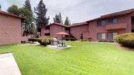 This residential unit is part of a well-maintained multi-building complex characterized by its distinctive terracotta-red and warm stucco exterior finish. The complex features a classic Southwestern-influenced architectural style, with textured stucco walls, low-pitched tile-accented rooflines, and sliding windows framed in neutral tones. The ground-floor unit is accessed via a covered concrete walkway, providing a sheltered and private entryway marked by a solid paneled door with exterior wall-mount lighting for added security and visibility.

The complex is set within lush, manicured grounds that serve as a central communal gathering space. A generous stretch of well-kept lawn anchors the outdoor area, complemented by mature evergreen trees that provide natural shade and privacy. A dedicated BBQ station with an outdoor grill, stone surround, and a picnic table shaded by a vibrant orange umbrella offers residents an inviting space for outdoor dining and socializing.

Exterior staircases with granite-textured treads and white metal railings provide clean, functional access to upper-level units. Trimmed hedgerows and ornamental grasses accent the building perimeters, adding greenery and definition to the walkways. The overall ambiance of the complex is practical, tidy, and community-oriented, offering a comfortable residential setting.