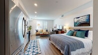 Located at 2525 Bayfront Blvd in Hercules, California, this modern studio presents a thoughtfully designed open-concept layout that seamlessly integrates the sleeping, living, and kitchen areas into one cohesive, light-filled space. Wide-plank wood-look flooring runs throughout, lending warmth and continuity to every zone.

The living area offers comfortable seating centered around a mid-century modern coffee table, with a large window dressed in wood-slat blinds that invites natural light and outdoor views. Adjacent to it, a dedicated workspace with a desk and ergonomic chair provides a functional nook for productivity.

The sleeping area features a generously sized bed with a tufted headboard, complemented by a wood media console and flatscreen TV. Tasteful coastal-inspired artwork adds character to the crisp white walls.

The kitchen is fully equipped with stainless steel appliances — including a refrigerator, electric range, built-in microwave, and dishwasher — set against white shaker cabinetry and a classic subway tile backsplash with quartz countertops.

The bathroom is clean and contemporary, showcasing a wide vanity with quartz countertop, a large mirror, ample storage, and a glass-enclosed walk-in shower. An in-unit washer is also present, adding everyday convenience to this well-appointed studio.