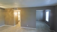 This single-story space presents a straightforward layout with carpeted flooring throughout, finished in a neutral gray and tan color palette. The main living area features a textured ceiling with a central HVAC vent, ensuring climate control throughout the space. The walls are painted in a cool gray tone, providing a calm, neutral backdrop. An open doorway to the right leads to an additional room, also carpeted, offering flexible use as a bedroom or den. To the left, a hallway connects to a bathroom, illuminated by a wall-mounted sconce that casts a warm, inviting glow. The bathroom area, visible through the hallway, appears to feature light-toned finishes. The layout is functional and open, with clear sightlines between the connected spaces. Electrical outlets are positioned along the baseboards for convenient access. White baseboards and trim provide a clean contrast to the gray walls. Overall, the space offers a simple, livable environment with a cohesive neutral design scheme suited for a variety of furnishing styles and uses.