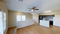 This two-story residence features an open-concept main living area that seamlessly connects the living room and kitchen, creating a spacious and functional layout. The flooring throughout the main level consists of warm-toned, hardwood-style planks that add a clean, contemporary feel to the space.

The living area is outfitted with a ceiling fan with integrated lighting, providing both airflow and ambient illumination. Recessed lighting supplements the natural light that filters in through multiple windows fitted with blinds, keeping the space bright and airy. A half-wall partition subtly defines the boundary between the living and kitchen areas while maintaining the open feel.

The kitchen is equipped with dark espresso-toned cabinetry that contrasts sharply with the lighter walls and flooring. A kitchen island with light stone-finish countertops offers additional workspace and casual seating potential. Stainless steel appliances, including a range and microwave, are integrated into the cabinetry. The neutral wall tones throughout create a versatile backdrop, while the overall design balances practicality with a modern aesthetic across both floors of the residence.