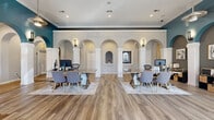 Located at 2299 River Oaks Dr in Columbus, Ohio, this single-floor leasing office and community welcome center makes a strong first impression with its refined architectural design. The grand entrance features double glass doors topped with a arched transom window, flanked by bold dark walls and white columns that set an elegant tone from the moment of arrival. Inside, the expansive open main area is defined by a series of stunning white arched colonnades, decorative wall niches, and teal accent walls that create a rich, layered aesthetic. Wood-look luxury flooring runs throughout, adding warmth and continuity across the space. Recessed lighting, pendant fixtures, and wall sconces provide a well-lit, welcoming atmosphere, while the "Welcome Home!" signage anchors the back wall as a focal point. Workstation areas are thoughtfully arranged on area rugs, giving the open floor plan a sense of defined purpose without sacrificing spaciousness. A separate dining area, accessed through an arched entry, features large windows that flood the space with natural light and offer pleasant outdoor views. This room includes seating and a wall-mounted display, making it suitable for meetings or resident gatherings. Overall, the space balances a professional, polished character with a warm, community-oriented ambiance.