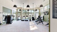 Located at 3650 Custer Pkwy in Richardson, TX, this fitness center offers a well-appointed, single-floor exercise space designed to support a comprehensive workout experience. The room is defined by soaring double-height ceilings and an expansive wall of green-trimmed windows that flood the interior with abundant natural light and provide views of the surrounding outdoors, including glimpses of a nearby pool area. The light-toned, patterned carpet flooring adds a refined aesthetic, while large wall mirrors along one side visually expand the space and support strength training focus. Overhead, a series of bold pendant lights complement ceiling fans, ensuring the room remains well-lit and comfortable. The cardio zone features multiple treadmills and elliptical machines, each equipped with individual display screens for an engaging workout. Strength training needs are addressed through a selection of cable and selectorized machines, a free-weight rack with dumbbells, and a flat bench. Green-painted architectural details, including framed doorways and wall paneling, lend the space a cohesive and polished look. Entryways with glass-paneled French doors provide a welcoming transition into the fitness area. This facility is thoughtfully designed to deliver both function and comfort for everyday fitness use.