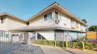 This multi-story residential building presents a classic Southern California architectural style, characterized by a smooth stucco exterior finished in a neutral cream tone with contrasting brown trim and a low-pitched roof. The property is accessed through a secured automatic sliding gate, providing a controlled entry point for residents and vehicles, with clearly posted caution signage emphasizing safety protocols.

The building's facade features a distinctive architectural detail — a circular arch design element set into a painted brick lower section, adding visual interest to the exterior. Wall-mounted light fixtures are positioned along the upper level, providing exterior illumination. Window-mounted air conditioning units are visible, servicing individual units throughout the building.

The landscaping along the front perimeter incorporates drought-tolerant plants, including agave and low shrubs, set within a gravel ground cover — a practical and low-maintenance design choice well-suited to the local climate. Yellow safety bollards line the entrance driveway, delineating the vehicle access path. A property management sign is prominently displayed near the street frontage, identifying the building's management affiliation. The overall setting reflects a well-maintained, functional residential community.