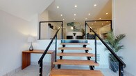 The Studios At Patchen showcases a thoughtfully designed interior defined by clean lines and modern aesthetics. A standout architectural feature is the open staircase with warm wood treads and sleek black steel cable railings, which serves as a visual centerpiece upon entry. The open layout connects living areas seamlessly, creating a sense of spaciousness and flow. Recessed lighting is installed throughout the ceiling, ensuring consistent, well-distributed illumination across the space. The interior palette is anchored by crisp white walls and light tile flooring, lending a bright, airy quality to the environment. A striking spiral staircase is visible in the background, adding a sculptural element that reinforces the contemporary design language. Carefully placed greenery introduces natural accents that complement the modern finishes. The overall ambiance is polished and inviting, balancing functional open-plan living with refined architectural details that make The Studios At Patchen a distinctive and well-appointed space.