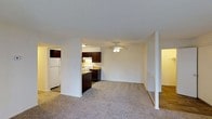 Located on Steinbeck Drive in Raleigh, North Carolina, this single-floor unit offers a practical and functional layout designed for comfortable everyday living. The open-concept design connects the living area and kitchen, creating a cohesive and accessible flow throughout the main space.

The living area is finished with wall-to-wall carpet, providing a soft and comfortable underfoot feel. A ceiling fan is centrally positioned, offering both light and air circulation to the space. The neutral white walls throughout contribute to a clean, bright aesthetic that allows for versatile decorating options.

The kitchen is semi-partitioned from the living space, featuring dark espresso-toned cabinetry that contrasts with the lighter surroundings. Under-cabinet lighting provides functional task illumination, and the kitchen includes essential appliances such as a white refrigerator and a range. A partial view reveals an adjacent hallway or secondary space, suggesting additional utility areas nearby.

Hardwood-style flooring is visible in a connected hallway area, offering a visual contrast to the carpeted living zone. The overall ambiance is straightforward and move-in ready, with a clean, unfussy interior well-suited to a variety of living arrangements.