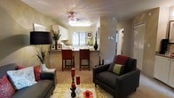 This single-story space showcases an open-concept layout that connects the living area and kitchen in a cohesive, functional design. The living room is anchored by plush, dark gray seating arranged around a white coffee table atop a patterned area rug, creating a comfortable and stylish gathering space. Warm green-toned walls contribute to the inviting ambiance throughout.

The kitchen is positioned at the rear of the open floor plan and features white cabinetry, a light countertop, and a breakfast bar with two rich, dark red bar stools — offering both additional seating and a natural division between the two spaces. A large window above the kitchen sink allows natural light to filter in, brightening the area. Framed artwork and decorative accents, including tall vases and potted plants, add personality and visual interest throughout.

Overhead, a ceiling fan with a light fixture provides both illumination and airflow. The flooring transitions from carpet in the living area to a hard surface in the kitchen, clearly delineating each functional zone while maintaining an open feel. A hallway visible to the right suggests additional rooms beyond the main living space.