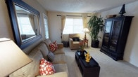 This single-floor residence offers a warm and inviting atmosphere anchored by a thoughtfully designed living room. The space is finished with plush wall-to-wall carpeting and neutral-toned walls that create a welcoming, comfortable environment throughout.

The living room serves as the heart of the home, bathed in natural light streaming through large windows dressed with elegant cream curtains and horizontal blinds. A textured ceiling adds dimension to the room, while a ceiling fan ensures year-round comfort. The seating area is generously sized, accommodating a plush tan sofa adorned with bold red and navy accent pillows, paired with a matching armchair. A sleek dark coffee table anchors the seating arrangement, complemented by decorative accents including gold fleur-de-lis figurines and a small floral arrangement.

A large statement mirror on one wall adds depth and light to the room, while a substantial dark wood entertainment unit provides ample storage and display space. A tall, lush indoor plant in the corner introduces a natural, organic element that enlivens the room. Warm ambient lighting from a classic table lamp rounds out the space, creating a cozy and functional living area suitable for both relaxing and entertaining.