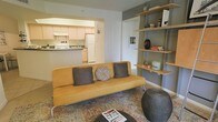 This single-floor residence offers a well-organized and stylish layout designed for comfortable everyday living. The living area serves as the heart of the space, anchored by a warm mustard-yellow sofa accented with decorative pillows and set atop a richly patterned area rug. A standout feature of the room is the built-in open shelving unit mounted along one wall, providing both storage and display space for books, plants, and personal décor. The neutral wall tones — a combination of soft white and cool gray — create a clean, modern backdrop that allows furnishings to take center stage. The kitchen is directly accessible from the living area through an open arched pass-through, creating a connected and airy feel between the two spaces. The kitchen features light wood cabinetry, white appliances, and a practical layout that maximizes functionality within a compact footprint. The overall ambiance of the space is casual yet curated, with a mix of modern and eclectic design elements that lend warmth and character throughout. Natural light filters into the living area, enhancing the inviting atmosphere of this well-appointed single-level home.