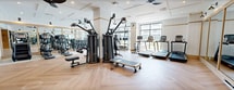 This 940-square-foot fitness center at 85 Livingston Ave in Roseland, NJ is a well-appointed, single-floor facility designed to deliver a comprehensive workout experience. The space features elegant herringbone-patterned hardwood flooring throughout, lending a refined aesthetic that sets it apart from a typical gym environment. Coffered white ceilings with recessed lighting and modern ceiling fans contribute to an open, airy atmosphere, while large floor-to-ceiling windows along multiple walls flood the space with natural light and offer views of the surrounding exterior.

The fitness center is equipped with a diverse range of commercial-grade equipment, including cable and multi-functional strength training machines, a free weight rack with dumbbells, adjustable benches, treadmills, ellipticals, stationary bikes, and a heavy punching bag with a functional training rig. Full-length gold-framed mirrors line one wall, visually expanding the space and supporting proper form during workouts.

A dedicated wellness station along the back wall features decorative tiling and built-in shelving, adding both functionality and visual interest. The entrance features sleek glass doors that connect seamlessly to the broader building. Wall-mounted accent lighting and clean white shiplap paneling complete the upscale, modern design of this thoughtfully curated fitness space.