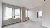 This single-floor unit is designed with a clean, neutral aesthetic, featuring light gray and beige tones across all rooms. The living area connects seamlessly to a pass-through kitchen equipped with light wood cabinetry, a range, and a built-in microwave, illuminated by recessed lighting above. A breakfast bar separates the kitchen from the carpeted living space, creating an open, functional layout.

The hallway connects the main living area to the bedrooms, featuring built-in countertop space along one wall and recessed lighting overhead. Multiple carpeted bedrooms are present, each featuring white horizontal blinds on windows that allow for adjustable natural light. One bedroom benefits from a corner window arrangement, bringing in light from two directions, while another includes dual mirrored bifold closet doors providing generous storage.

Two bathrooms are included in the unit. One features a white vanity with a medicine cabinet above, while the second offers a similarly clean design with tile flooring, a white vanity, mirror, and chrome fixtures. Both are finished in crisp white tones. Throughout the unit, white baseboards, smooth ceilings, and consistent carpeting contribute to a cohesive and move-in ready presentation.