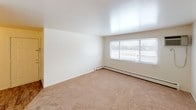 This single-floor residential unit offers a practical and well-organized layout across several distinct spaces. The living area is generously sized, featuring warm beige carpeting, clean white walls, and a baseboard heating system beneath a large window that allows natural light to fill the room. The open sightlines from the living area extend toward the kitchen, creating a connected and functional flow throughout the unit.

The galley-style kitchen is efficiently designed with dark wood cabinetry lining both walls, offering ample storage space. Light-toned countertops provide a contrast to the cabinetry, while a double-basin sink, a white gas range, and a full-size white refrigerator complete the functional setup. Hardwood-style flooring and overhead lighting give the kitchen a clean, practical feel.

Two carpeted bedrooms provide comfortable personal spaces, each featuring baseboard heating and windows fitted with horizontal blinds that offer both natural light and privacy. The rooms are bright and simply finished with white walls and neutral flooring.

The bathroom is clean and straightforward, featuring a full-size tub and shower combination with a white surround, a toilet, and a vanity, all set on hardwood-style flooring. The overall aesthetic of the unit is neutral, functional, and move-in ready.