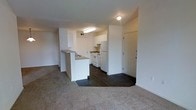 This single-floor space features an open-concept design that seamlessly connects the living area and kitchen. The living area is finished with neutral-toned carpet, providing a comfortable and understated foundation, while the kitchen and entryway transition to dark hardwood-style flooring, creating a clear visual distinction between spaces.

The kitchen is equipped with white cabinetry, a white refrigerator, and a white electric range with overhead ventilation. A peninsula counter extends into the open layout, offering additional workspace and serving as a natural divider between the kitchen and the main living area. Upper cabinets line the kitchen walls, providing ample storage.

The entryway, positioned adjacent to the kitchen, features a solid white door and is clearly defined within the floor plan. White walls throughout the space contribute to a bright, clean atmosphere, complemented by overhead flush-mount and fluorescent lighting. A corridor visible to the left suggests access to additional rooms. The overall design is straightforward and functional, with a neutral palette that offers versatility for personalization.