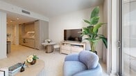 Located on the 38th floor at 77 SE 5th St in Miami's vibrant Brickell neighborhood, this well-appointed unit offers a thoughtfully designed single-floor layout encompassing two living areas, two bathrooms, a hallway, and a private balcony. The interior presents a cohesive, modern aesthetic defined by a soft neutral palette of warm whites and natural wood tones, complemented by light tile flooring throughout.

The primary living area is bright and welcoming, featuring a plush seating arrangement, a built-in workspace with a light wood desk, and a pair of abstract teal-toned artworks that add a pop of color to the serene backdrop. A second living space opens to a compact dining area and a kitchen fitted with light wood cabinetry, maintaining the unit's consistent warm tone. Large sliding glass doors lead to the private balcony, where a glass-panel railing frames open city views of the Brickell skyline.

Both bathrooms are finished with stone-look tile walls and floors, featuring soaking tub and shower combinations with brushed gold fixtures, offering a refined, spa-like atmosphere. A dedicated walk-in closet with custom light wood shelving and hanging space provides generous storage. Natural light flows throughout, enhancing the clean, contemporary feel of this well-planned Miami residence.