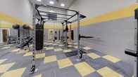 This fitness facility is designed to support a comprehensive range of training styles, from strength and resistance work to functional and combat-inspired exercise. The space is immediately recognizable by its striking interior design: walls are finished in a two-tone gray and yellow horizontal band pattern, which is echoed by the checkered yellow and gray rubber flooring below — a cohesive aesthetic that is both energetic and practical.

The facility is furnished with an impressive array of equipment. A large multi-function power rack serves as a central feature, supporting TRX suspension trainers, pull-up bars, and punching bags, including a full-size Century heavy bag. Adjacent to the rack, a wall-mounted horizontal bar system provides additional pull and hang training options. The space also includes multiple adjustable weight benches, a dedicated cable machine, a barbell with a full plate storage rack, and an assortment of dumbbells and kettlebells. Medicine balls and stability balls are neatly organized on a dedicated storage rack.

A large wall mirror expands the sense of space and allows users to monitor their form during training. Recessed ceiling lights provide bright, even illumination throughout. The layout is open and logically arranged, offering ample room to move freely between stations, making this facility well-suited for individual workouts or small group training sessions.