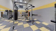 This fitness facility is designed to support a comprehensive range of training styles, from strength and resistance work to functional and combat-inspired exercise. The space is immediately recognizable by its striking interior design: walls are finished in a two-tone gray and yellow horizontal band pattern, which is echoed by the checkered yellow and gray rubber flooring below — a cohesive aesthetic that is both energetic and practical.

The facility is furnished with an impressive array of equipment. A large multi-function power rack serves as a central feature, supporting TRX suspension trainers, pull-up bars, and punching bags, including a full-size Century heavy bag. Adjacent to the rack, a wall-mounted horizontal bar system provides additional pull and hang training options. The space also includes multiple adjustable weight benches, a dedicated cable machine, a barbell with a full plate storage rack, and an assortment of dumbbells and kettlebells. Medicine balls and stability balls are neatly organized on a dedicated storage rack.

A large wall mirror expands the sense of space and allows users to monitor their form during training. Recessed ceiling lights provide bright, even illumination throughout. The layout is open and logically arranged, offering ample room to move freely between stations, making this facility well-suited for individual workouts or small group training sessions.