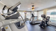 This fitness center is a well-appointed exercise room designed to accommodate a variety of cardio-focused workouts. The space features durable dark rubber flooring throughout, providing a practical and resilient surface suited to heavy equipment use. Crown molding and a textured ceiling add a degree of architectural refinement, while a ceiling fan with integrated lighting ensures comfortable air circulation during workouts.

Multiple windows fitted with horizontal blinds line the walls, allowing abundant natural light to fill the room and offering views of the surrounding outdoor environment. This windowed design creates an open, airy atmosphere despite the room's compact footprint. A wall-mounted flat-screen television is positioned for visibility from several exercise stations, enhancing the workout experience.

The equipment lineup includes treadmills, elliptical cross-trainers, a stair climber, and a stationary recumbent bike, representing brands such as Cybex and True Fitness. A weight bench and additional strength accessories are also present. Entry is provided through a set of elegant French doors, contributing a classic design touch. A wall mirror in one section visually expands the space, reinforcing its functional and welcoming character.