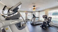 This fitness center is a well-appointed exercise room designed to accommodate a variety of cardio-focused workouts. The space features durable dark rubber flooring throughout, providing a practical and resilient surface suited to heavy equipment use. Crown molding and a textured ceiling add a degree of architectural refinement, while a ceiling fan with integrated lighting ensures comfortable air circulation during workouts.

Multiple windows fitted with horizontal blinds line the walls, allowing abundant natural light to fill the room and offering views of the surrounding outdoor environment. This windowed design creates an open, airy atmosphere despite the room's compact footprint. A wall-mounted flat-screen television is positioned for visibility from several exercise stations, enhancing the workout experience.

The equipment lineup includes treadmills, elliptical cross-trainers, a stair climber, and a stationary recumbent bike, representing brands such as Cybex and True Fitness. A weight bench and additional strength accessories are also present. Entry is provided through a set of elegant French doors, contributing a classic design touch. A wall mirror in one section visually expands the space, reinforcing its functional and welcoming character.