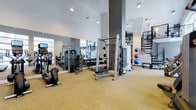 This well-appointed, two-story fitness center offers a spacious and thoughtfully designed environment for a thorough workout experience. The ground floor is outfitted with a diverse selection of exercise equipment, including a Smith machine, cable machines, a dumbbell rack, and cardio options such as a recumbent bike. Stability balls and additional accessories are neatly organized along the walls, keeping the floor space open and easy to navigate. Large mirrors line the walls, amplifying the sense of space and allowing users to monitor their form during training. The flooring features a neutral, textured carpet tile that adds comfort underfoot throughout the workout area. Soaring ceilings with pendant lighting contribute to an airy, open atmosphere, while natural light filters in through large windows, creating a bright and energizing environment. A sleek, modern spiral staircase connects the two levels, serving as both a functional and architectural focal point. Structural columns punctuate the space, complementing the contemporary aesthetic. The second floor is accessible via the spiral staircase, expanding the available workout and amenity space. Overall, the fitness center combines modern design with practical functionality, making it a well-rounded facility suited for a variety of fitness routines.