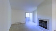 This single-floor residence offers a clean, functional layout designed for comfortable everyday living. The living area serves as the central focus of the space, characterized by neutral white walls, smooth ceilings, and wall-to-wall carpeting that lends a warm, understated feel underfoot. A standout feature of the room is the tile-surround fireplace, framed in dark and light contrasting tile work with a raised hearth, adding both a practical heating element and a visual focal point to the space. Natural light flows generously into the living area through a full-height sliding glass door dressed with vertical blinds, offering access to the outdoors while brightening the interior throughout the day. A baseboard heating unit runs along the exterior wall beneath the sliding door, ensuring efficient climate control throughout the cooler months. The overall ambiance of the space is clean and minimalist, presenting a neutral canvas well-suited for a variety of interior styles and furnishings. The single-floor configuration provides ease of movement and accessibility throughout, making it a practical and inviting living environment.