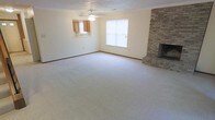 This two-floor home offers a thoughtfully laid-out living space with practical, classic design elements throughout. The main living area is generously sized, finished with wall-to-wall carpet that adds warmth underfoot. A standout feature of the room is the floor-to-ceiling brick fireplace positioned in the corner, serving as a natural focal point and adding a traditional, cozy character to the space. A ceiling fan overhead provides both lighting and ventilation, contributing to the room's comfort year-round. Natural light enters through a centrally placed window fitted with blinds, keeping the pale, neutral-toned walls bright and inviting. A pass-through window opens directly into the kitchen area, where wooden cabinetry is visible, offering a connected, open feel between the living and kitchen spaces while maintaining distinct zones. The textured ceiling adds a subtle layer of dimension to the room's finish. The overall ambiance is clean, understated, and functional, providing a solid foundation for comfortable everyday living across its two floors.