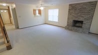 This two-floor home offers a thoughtfully laid-out living space with practical, classic design elements throughout. The main living area is generously sized, finished with wall-to-wall carpet that adds warmth underfoot. A standout feature of the room is the floor-to-ceiling brick fireplace positioned in the corner, serving as a natural focal point and adding a traditional, cozy character to the space. A ceiling fan overhead provides both lighting and ventilation, contributing to the room's comfort year-round. Natural light enters through a centrally placed window fitted with blinds, keeping the pale, neutral-toned walls bright and inviting. A pass-through window opens directly into the kitchen area, where wooden cabinetry is visible, offering a connected, open feel between the living and kitchen spaces while maintaining distinct zones. The textured ceiling adds a subtle layer of dimension to the room's finish. The overall ambiance is clean, understated, and functional, providing a solid foundation for comfortable everyday living across its two floors.