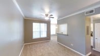 This single-floor space offers a clean, functional layout centered around a carpeted open living area. The room is finished with white crown molding that frames the ceiling, adding a polished architectural detail against the warm taupe-toned walls. A ceiling fan with a built-in light fixture is centrally positioned, providing both comfort and illumination. Natural light enters through a large window on the far wall, offering a view of lush greenery outside and brightening the interior with a welcoming glow. A second window in the adjacent dining area further enhances the sense of openness. The living and dining areas flow seamlessly together, creating a cohesive shared space. A partial wall with a pass-through granite-topped counter connects to the kitchen, which is visible in the background and features a chandelier-style light fixture, hinting at an elegant touch in a practical space. The neutral color palette throughout—warm gray walls, white trim, and beige carpet—creates a versatile backdrop suitable for a variety of interior styles. Overall, the space presents a comfortable, well-appointed layout with tasteful finishes.