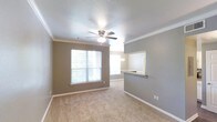 This single-floor space offers a clean, functional layout centered around a carpeted open living area. The room is finished with white crown molding that frames the ceiling, adding a polished architectural detail against the warm taupe-toned walls. A ceiling fan with a built-in light fixture is centrally positioned, providing both comfort and illumination. Natural light enters through a large window on the far wall, offering a view of lush greenery outside and brightening the interior with a welcoming glow. A second window in the adjacent dining area further enhances the sense of openness. The living and dining areas flow seamlessly together, creating a cohesive shared space. A partial wall with a pass-through granite-topped counter connects to the kitchen, which is visible in the background and features a chandelier-style light fixture, hinting at an elegant touch in a practical space. The neutral color palette throughout—warm gray walls, white trim, and beige carpet—creates a versatile backdrop suitable for a variety of interior styles. Overall, the space presents a comfortable, well-appointed layout with tasteful finishes.
