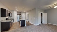 This single-floor residence offers a thoughtfully designed open-concept layout that seamlessly connects the kitchen and living areas. The kitchen is a standout feature, showcasing rich dark-stained cabinetry, light granite countertops, and a full suite of stainless steel appliances including a refrigerator, range, and over-the-range microwave. A central island with a built-in sink and pendant lighting above serves as both a functional workspace and a natural gathering point. Wood-look flooring runs throughout the kitchen, transitioning to plush carpet in the spacious living area. The living space is generously proportioned, featuring neutral gray walls, a ceiling fan with light fixture, and a sliding glass door fitted with vertical blinds that allows natural light to fill the room. Multiple interior doors provide access to additional rooms and closet storage, including what appears to be a walk-in closet. The bathroom features a clean, all-white tub and shower combination with chrome fixtures, set against warm wood-look flooring and soft gray walls. Recessed lighting throughout the home, along with clean white trim and baseboards, gives the space a modern, polished aesthetic.