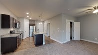 This single-floor residence offers a thoughtfully designed open-concept layout that seamlessly connects the kitchen and living areas. The kitchen is a standout feature, showcasing rich dark-stained cabinetry, light granite countertops, and a full suite of stainless steel appliances including a refrigerator, range, and over-the-range microwave. A central island with a built-in sink and pendant lighting above serves as both a functional workspace and a natural gathering point. Wood-look flooring runs throughout the kitchen, transitioning to plush carpet in the spacious living area. The living space is generously proportioned, featuring neutral gray walls, a ceiling fan with light fixture, and a sliding glass door fitted with vertical blinds that allows natural light to fill the room. Multiple interior doors provide access to additional rooms and closet storage, including what appears to be a walk-in closet. The bathroom features a clean, all-white tub and shower combination with chrome fixtures, set against warm wood-look flooring and soft gray walls. Recessed lighting throughout the home, along with clean white trim and baseboards, gives the space a modern, polished aesthetic.