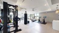 Located in Miami, FL, this well-appointed fitness center offers a spacious, single-floor layout designed to accommodate a full spectrum of workout needs. The space is defined by its open, airy design, with floor-to-ceiling windows and large glass doors lining multiple walls, flooding the interior with abundant natural light and providing views of the surrounding greenery and outdoor areas.

The cardio zone is comprehensively equipped with treadmills, elliptical machines, Assault fitness runners, and stationary cycling bikes featuring interactive screens. A dedicated strength training area houses a full dumbbell rack, an Octagon functional training rig, battle ropes, a heavy punching bag, and plyo boxes, supporting both high-intensity and traditional resistance training. Full-length mirrors along one wall enhance the sense of space and allow for form monitoring during workouts.

The flooring throughout combines light-toned, textured fitness flooring with rubber mat zones beneath equipment, ensuring both safety and durability. Ceiling fans provide additional ventilation, complementing the bright, recessed lighting overhead. A refreshment station with a mini-refrigerator and storage is tucked into one corner, adding convenience. Large glass doors open directly to an outdoor turf area, seamlessly extending the workout experience beyond the walls of the facility.