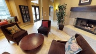 This single-floor space presents a warm and well-appointed lobby area designed for comfort and community. The interior is anchored by a generous seating arrangement featuring plush dark upholstered armchairs accented with rich red and patterned throw pillows, gathered around a rounded wooden coffee table — creating an inviting lounge atmosphere. The floors are finished in large-format light travertine tiles that lend an elegant, airy feel to the space.

A tall, glass-enclosed media or library cabinet lines one wall, offering an extensive collection that adds both function and visual interest. Adjacent to the seating area, a substantial stone fireplace provides a classic focal point and contributes to the overall warmth of the environment. A large, lush potted tree adds a natural, organic element that softens the architectural lines and enhances the welcoming ambiance.

The space benefits from soaring ceilings and abundant natural light streaming through full-length glass entry doors, creating a bright and open feel. Warm golden-yellow walls complement the neutral tile floors and rich wood tones throughout. Wall sconces provide soft supplemental lighting, adding to the refined yet approachable character of this thoughtfully designed communal space.