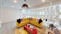 This single-floor common area is a lively, well-appointed space that blends modern design with mid-century flair. The main lounge area is anchored by a bold mustard-yellow tufted sectional sofa, accessorized with an eclectic mix of textured throw pillows, and set atop a striped yellow and tan area rug. A striking red lacquered coffee table serves as a vibrant focal point, stacked with reading materials for guests. Overhead, a large dome pendant light in brushed bronze adds an industrial-chic touch to the bright white ceiling with recessed lighting throughout.

A defining architectural feature is the floor-to-ceiling open-cube bookshelf that acts as a stylish room divider, allowing natural light to pass through while subtly separating the lounge from adjacent spaces. Large windows and glass doors flood the interior with daylight and provide clear views to an outdoor patio area furnished with colorful seating.

Beyond the lounge, a corridor-style media area features wall-mounted flat-screen televisions, light wood paneling, and sleek white cabinetry, creating a functional yet polished entertainment zone. Neutral large-format tile flooring runs throughout, providing a clean, cohesive foundation that complements the space's vibrant color palette. The overall ambiance is energetic, welcoming, and thoughtfully designed for social interaction.
