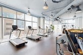 Located at 60 11th St NE in Atlanta's Midtown neighborhood, this expansive fitness facility spans a single floor and comprises two dedicated exercise rooms designed to support a wide range of workout disciplines. The main exercise area is defined by dramatic floor-to-ceiling windows that flood the space with natural light and frame sweeping views of the Atlanta skyline, creating an energizing atmosphere for cardio training. Rows of treadmills, ellipticals, stationary bikes, and rowing machines are complemented by a comprehensive free weights section featuring a full dumbbell rack, barbells, and Matrix-brand strength machines. Exposed ductwork runs overhead in a structured industrial style, while pendant lights provide focused illumination throughout. Durable rubber flooring covers the workout areas, offering both comfort and safety. The second exercise room serves as a dedicated studio space, outfitted with spin bikes, wall-mounted mirrors, exercise mats, kettlebells, and a flat-screen display for guided workouts. This room features a clean, minimalist aesthetic with tile flooring and bright linear lighting. Together, the two rooms provide a comprehensive, well-organized fitness environment suited for both individual training and group exercise activities.