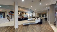 This sophisticated single-floor community space is designed for social engagement and relaxation, blending contemporary architecture with warm, inviting interiors. The expansive open-plan layout features a seamless transition between distinct zones, unified by a mix of wood-look flooring and large-format tile inlaid with a decorative mosaic border. A dramatic curved tray ceiling with recessed lighting anchors the central lounge, where a sweeping, curved sectional sofa in cream upholstery serves as an eye-catching focal point. Adjacent to the lounge is a stylish bar area appointed with a rich wood-topped counter, bar-height seating, pendant lighting, and a mirrored back bar display. Steps with decorative iron railings lead to and from this zone, adding architectural depth to the layout. A games area accommodates a full-size billiards table alongside additional game tables, all beneath a bold circular tray ceiling. The TV lounge offers an intimate retreat, featuring built-in wall niches with artistic displays, a wall-mounted flat-screen TV, and curated seating on a patterned area rug. At one end, a grand atrium-style entry soars with double-height ceilings and large windows that flood the space with natural light, completed with plush seating arrangements and colorful artwork throughout.