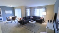 This single-floor residence features an open-concept living area filled with abundant natural light streaming through multiple large windows dressed with patterned curtains and horizontal blinds. The living room is anchored by a generous L-shaped navy sectional sofa accented with coral and patterned throw pillows, positioned around a modern white round coffee table atop a chevron-patterned area rug over neutral wall-to-wall carpeting. A sleek floor lamp and side tables with complementary lighting add warmth to the space. The walls are finished in a soft gray tone, contributing to the room's calm, contemporary aesthetic. Sliding glass doors along one wall suggest access to an outdoor area, enhancing the sense of openness and connectivity to the exterior. A dining area is partially visible to the side, featuring a small table and chairs, indicating a practical separation of living and dining functions within the open layout. Overall, the space presents a cohesive, modern design with a neutral palette, ample natural light, and thoughtfully arranged furnishings suited for comfortable everyday living.