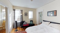 This single-floor space is a well-utilized studio-style room that combines sleeping and lounge areas into one cohesive living space. The room is finished with warm hardwood floors and cream-colored walls, creating a bright and inviting atmosphere. Two windows dressed with sheer white curtains and gray drapes allow generous natural light to filter in, enhancing the airy feel of the space. A classic cast-iron radiator beneath one window underscores the building's traditional character.

The sleeping area features a generously sized bed dressed in crisp white linens, positioned to make efficient use of the available floor space. A black futon sofa with decorative throw pillows serves as a sitting area, adding versatility to the room. A traditional patterned area rug anchors the space, providing a layer of texture and color contrast against the hardwood floors. An arc floor lamp offers additional ambient lighting.

A doorway visible from the main room leads to an en-suite bathroom, adding a level of privacy and convenience. The overall layout is practical and comfortable, suited for everyday living.