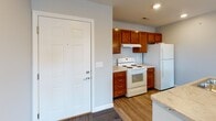 Located at 405 E Sycamore St in Kokomo, Indiana, this single-floor unit offers a practical and well-appointed layout designed for comfortable everyday living. The kitchen serves as a central feature, showcasing warm-toned wood cabinetry, light stone-look countertops, and a peninsula with a built-in sink that opens toward the living area. Decorative glass pendant lights hang above the peninsula, adding a refined touch, while recessed lighting and white appliances—including an electric range and refrigerator—complete the functional space. Hard surface flooring in the kitchen transitions to patterned carpet tiles throughout the living and bedroom areas, providing a cohesive yet defined flow between spaces. The living area is generously proportioned, featuring neutral gray walls, recessed lighting, and a glass-paned exterior door that invites natural light and outdoor views. The bedrooms are carpeted and well-lit by windows overlooking the surrounding neighborhood. The bathroom includes a wood-toned vanity with a white countertop, a large mirror with a lighted fixture above, and a full tub-shower combination, all finished with wood-look flooring. Throughout the unit, clean lines and neutral tones create a bright, move-in-ready atmosphere.