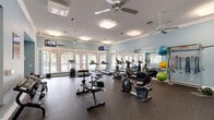 Located at 25455 Borough Park Dr in Spring, TX, this fitness center offers a comprehensive workout environment within a generously sized, single-level space. The room is finished with soft blue walls, crisp white crown molding, and durable rubber flooring, creating a clean and energizing atmosphere. Multiple ceiling fans and flush-mounted light fixtures ensure consistent airflow and bright, even illumination throughout.

Large white-framed windows and glass-paned doors line the perimeter, flooding the interior with abundant natural light and offering views of the surrounding outdoor greenery. A full-length wall mirror along one side visually expands the space and supports proper exercise form.

The fitness center is thoroughly equipped for a wide range of workouts. Cardio equipment includes treadmills, elliptical trainers, stationary bikes, and a stair climber. A dedicated free-weight area features a tiered dumbbell rack and flat benches, while a strength and functional training zone offers medicine balls, stability balls, resistance bands, and a TRX suspension system. Mounted televisions are positioned for convenient viewing during cardio sessions. The open floor plan allows for fluid movement between workout zones, making this fitness center a well-rounded facility suited to a variety of fitness routines and experience levels.