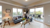 This single-floor residence offers a contemporary, open layout designed to maximize natural light and urban views. The living room serves as the centerpiece of the unit, featuring oversized windows on multiple walls and a sliding glass door that opens onto a private balcony. The outdoor space overlooks a lush, tree-lined neighborhood with a city skyline visible in the distance, creating a compelling indoor-outdoor connection. Light wood-tone flooring flows consistently throughout the unit.

The kitchen is well-equipped with flat-front cabinetry in a light wood finish, stainless steel appliances including a full-size refrigerator and electric range, a stainless steel wall-mounted range hood, and a chevron-patterned tile backsplash. A central island with a quartz-style countertop provides additional workspace and casual dining.

Two bedrooms each feature large windows with neighborhood views and dedicated closet space. The primary bedroom includes a tray ceiling detail, adding architectural interest. Two full bathrooms serve the unit; one includes a glass-enclosed walk-in shower, and both feature wood-look tile flooring, white vanities, and neutral wall tones. The overall aesthetic is clean and modern throughout.