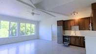 This single-floor space is defined by its clean, minimalist design and open-plan layout that seamlessly integrates the living area with the kitchen. The vaulted ceiling with exposed beams adds a sense of height and architectural character, while a large ceiling fan enhances both form and function. A generous picture window along one wall frames lush greenery outside, bathing the interior in abundant natural light and creating a strong connection to the outdoors. The polished concrete flooring extends throughout, providing a sleek, durable, and cohesive foundation that complements the contemporary aesthetic. The kitchen is equipped with rich, warm-toned wood cabinetry paired with light stone countertops, offering a classic contrast to the otherwise modern surroundings. Stainless steel appliances, including a full-size refrigerator and a dishwasher, ensure the kitchen is fully functional and ready for use. Recessed track lighting above the kitchen provides focused illumination. The open living area offers generous space, keeping the overall atmosphere airy and uncluttered. A doorway leads to an additional room, suggesting further utility within the layout. This space presents a well-balanced blend of practical design and modern finishes throughout.
