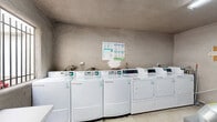 Located at 3500 Cedar Ave in Long Beach, California, this single-floor utility room serves as a functional shared service space. The room is finished with textured stucco walls and light tile flooring throughout, giving it a practical, no-frills character suited to its purpose.

The laundry area is equipped with multiple commercial-grade front-load washing machines and dryers from Speed Queen and WASH, arranged along the walls for efficient use of the available floor space. Usage instructions are posted on the wall for resident reference. A large metallic exhaust duct is prominently visible, venting the dryer units appropriately.

A dedicated mechanical alcove houses the building's water heating infrastructure, including multiple water heaters connected by copper piping and insulated ductwork. Safety signage is posted on the equipment, and a fire alarm panel is mounted on the adjacent wall.

Near the entry, a barred window allows natural light to filter into the space, offering a view of the surrounding outdoor courtyard with visible greenery and the building exterior. A red metal door provides direct access to the exterior. A folding table is positioned near the window, offering a surface for sorting or folding laundry. A fire extinguisher cabinet and cleaning supplies are also present, rounding out the room's practical amenities.