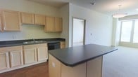 This single-floor space offers a functional and open layout, with the kitchen serving as a central feature. The kitchen is equipped with light wood cabinetry that provides ample storage, complemented by dark countertop surfaces that create a subtle contrast. A built-in sink is positioned along the perimeter counter, accompanied by a dishwasher and additional lower cabinetry for practical storage. A kitchen island with a matching dark countertop anchors the space, offering extra workspace and a natural dividing point between the kitchen and the adjacent living area. The flooring transitions from wood-style flooring in the kitchen to carpet in the living area, distinguishing the two zones while maintaining an open-concept feel. Neutral-toned walls throughout contribute to a clean and understated aesthetic. A doorway visible from the main area leads to an additional room, suggesting versatile use of space. The layout is straightforward and practical, designed to maximize usability within a compact footprint while maintaining an open and connected flow between the kitchen and living areas.