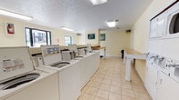Located at 2414 Stone Rd in East Point, Georgia, this single-floor laundry care center offers a practical and well-organized space designed to meet residents' laundry needs efficiently. The facility is finished with light beige tile flooring throughout, contributing to a clean and easy-to-maintain environment. Walls are painted in a soft, neutral yellow tone, lending the space a bright and welcoming atmosphere.

The main laundry area features a row of top-loading washing machines positioned along a central bank, complemented by stacked washer-dryer units along the wall, providing ample laundry capacity. Generous granite-topped folding counters run along both sides of the room, offering plenty of surface area for sorting and folding laundry. Overhead fluorescent lighting ensures the entire space is well illuminated.

An open corridor connects the main laundry area to the entrance, with large windows along one side allowing natural light to filter in. A clearly posted "Clothes Care Center" sign with usage guidelines is visible throughout the space, keeping users informed of proper procedures. A fire extinguisher is conveniently mounted near the entry for safety. The overall layout is straightforward and functional, making this laundry facility a convenient and accessible amenity.