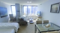 This well-appointed single-floor studio offers an efficient and thoughtfully arranged open-plan layout that combines living, sleeping, and dining areas into one cohesive space. Large floor-to-ceiling windows dominate the exterior wall, framing dramatic panoramic views of a dense urban skyline and allowing abundant natural light to pour in throughout the day, creating a bright and airy atmosphere.

The living area features a plush gray sofa and armchair arranged around a glass-and-metal coffee table, providing a comfortable seating arrangement with the skyline as a backdrop. A media console with a flat-screen television is positioned beneath the windows, making full use of the view as an ambient feature. Decorative glass bottles and subtle greenery add refined touches to the space.

The sleeping area is integrated seamlessly to one side, anchored by a neatly made bed with crisp white linens. A glass dining table with upholstered chairs rounds out the space, offering a dedicated area for meals or work. The neutral color palette—soft grays, whites, and warm tones—combined with carpeted flooring contributes to a calm, cohesive aesthetic. Overall, the space delivers a modern, urban living experience with a strong emphasis on natural light and city views.