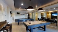 This single-floor recreational space is thoughtfully designed to offer a premium leisure experience with a seamless blend of modern style and functional layout. The centerpiece of the room is a full-size billiards table finished in a rustic wood frame with a neutral felt surface, complemented by a second pool table nearby, making it an ideal setting for entertainment and social gatherings. Overhead, striking dark dome pendant lights hang from a tray ceiling painted in deep charcoal, adding a dramatic contrast to the light hardwood flooring below. Recessed lighting throughout further enhances the ambiance. Along one wall, a sleek wet bar area features dark cabinetry, open shelving with decorative accents, and a utility sink, providing a convenient refreshment station. A cue rack is mounted nearby, keeping the space organized and game-ready. Glass partition walls and sliding doors connect the billiards area to an adjacent dining space, which is furnished with a dark table and upholstered chairs, offering a more intimate setting. The overall design is cohesive and contemporary, with clean lines, dark tones, and quality materials combining to create a polished and inviting entertainment environment.