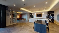 The Magnolia Lounge is a thoughtfully designed single-floor amenity space that seamlessly blends social, dining, and recreational functions into one cohesive environment. The space is divided into distinct yet connected zones, each with its own character and purpose.

The lounge area features warm hardwood-style flooring, a striking tray ceiling with a golden wood inlay, and a bold wall-mounted flat-screen TV set against a dark geometric accent wall. Vibrant terracotta-toned lounge chairs surround a sleek round coffee table, while an oversized pendant light anchors the space from above. Floor-to-ceiling windows flood the area with natural light and offer views of the surrounding exterior.

Adjacent to the lounge, a well-appointed kitchen and dining area is equipped with clean white quartz countertops, warm wood cabinetry, dual beverage refrigerators, a sink, and a large central island with seating. Globe pendant lights hang above the island, and bold circular artwork adorns the walls, adding an artistic focal point.

A slatted white room divider elegantly separates the entertaining areas from a dedicated game room, which houses a full-size billiards table set atop a patterned area rug beneath a low-hanging pendant light. Large-format tile flooring runs throughout, unifying the space. Recessed lighting and ambient LED cove details add warmth and depth throughout the entire lounge.